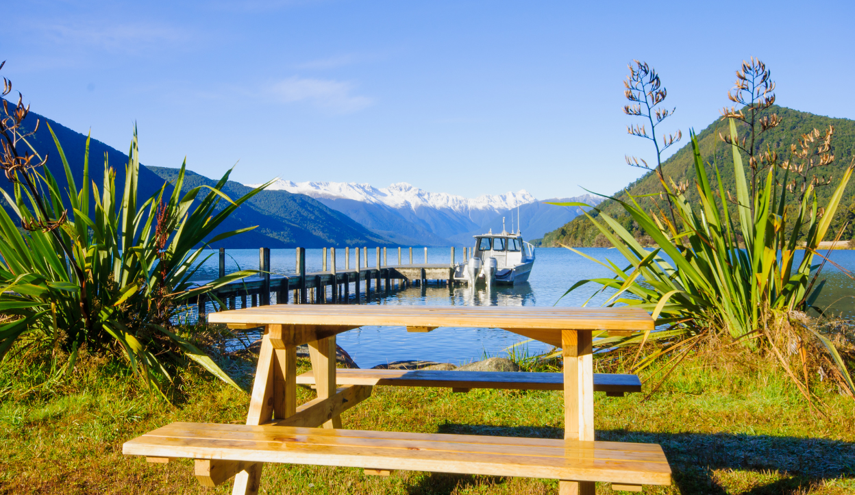 Wooden picnic table and dock by a clear alpine lake at Nelson Lakes National Park, New Zealand, with snow-capped mountains in the distance.