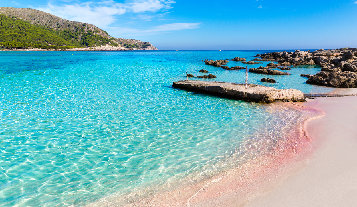 Crystal-clear water and soft white sand at Cala Agulla Beach in Mallorca, Spain, with rocky outcrops and forested hills in the background.