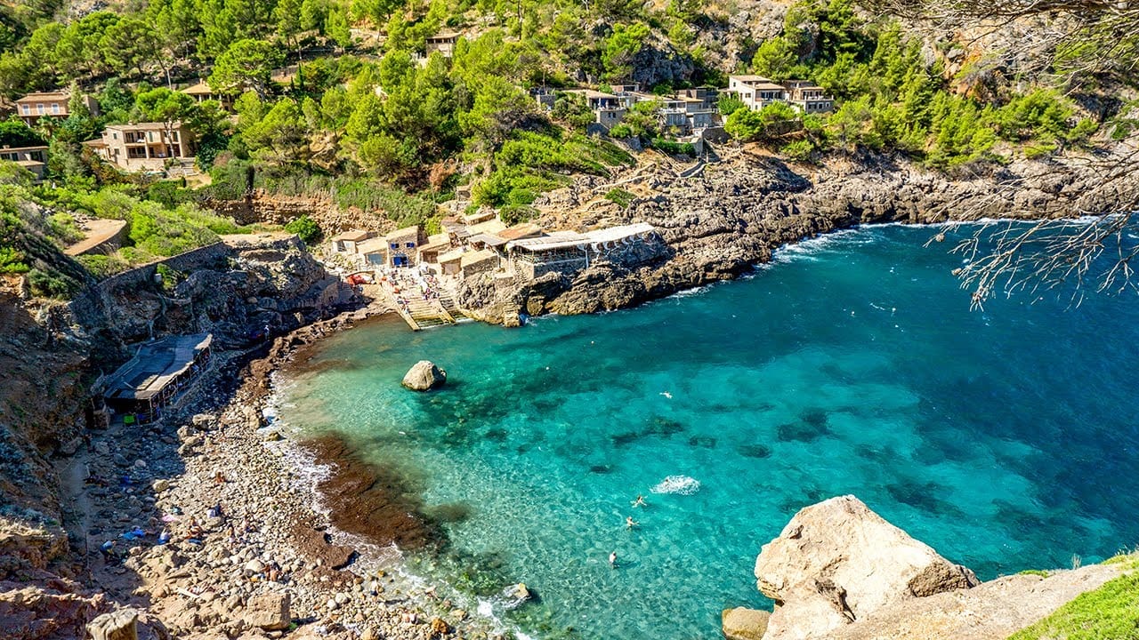 Rocky beach at Cala Deià in Mallorca, Spain, with clear water, traditional stone buildings, and forested cliffs.