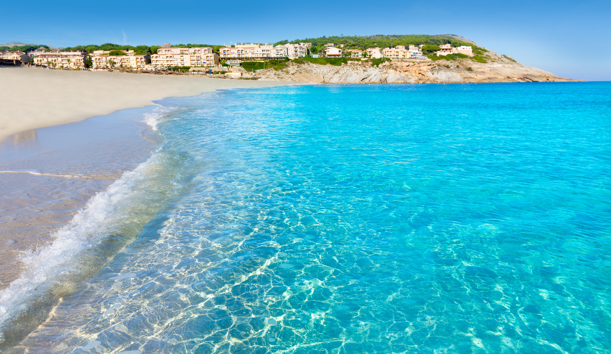 Shallow turquoise water gently washing onto the soft sandy shore at Cala Mesquida Beach in Mallorca, Spain, with seaside resorts on a rocky hillside in the background.