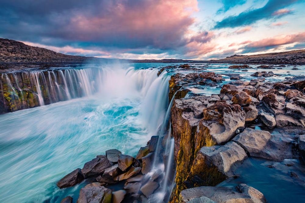 Dettifoss waterfall in northern Iceland captured at sunset, with powerful glacial water plunging over rugged basalt cliffs and swirling into a wide canyon under a colorful sky.