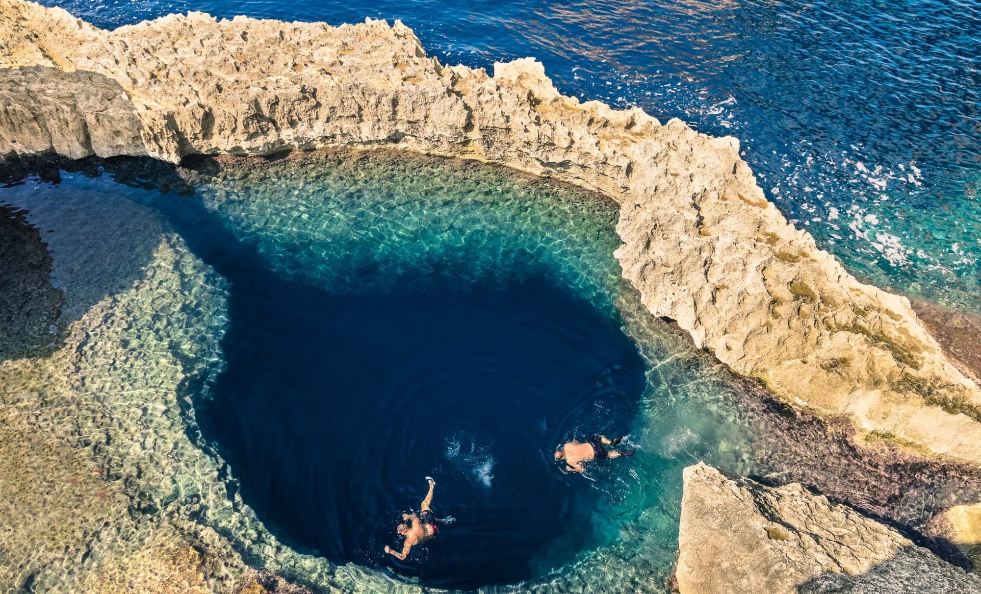 Overhead view of Dwejra Bay in Gozo, Malta, showing a deep blue natural pool with two snorkelers surrounded by rugged limestone rocks and clear shallow waters.