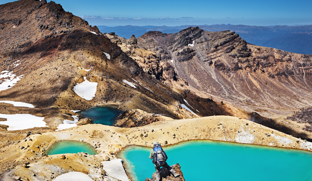Hiker overlooking vivid emerald lakes surrounded by volcanic terrain and rugged peaks in Tongariro National Park, New Zealand.