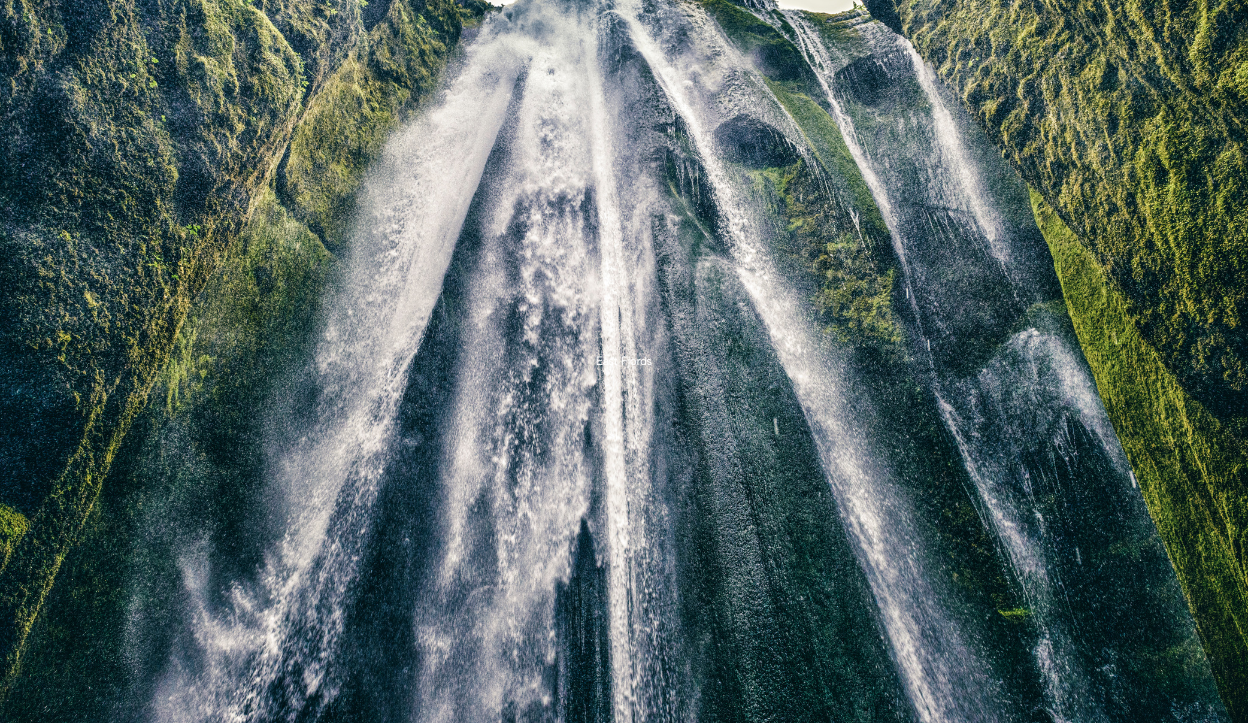 Upward view of Gljúfrabúi waterfall flowing through a moss-covered cliff crevice in southern Iceland, with water mist and dramatic light filtering through the canyon walls.