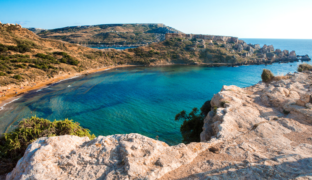 View from a rocky cliff overlooking Golden Bay in Malta, with a sandy beach and turquoise waters framed by rugged coastline and rolling hills.
