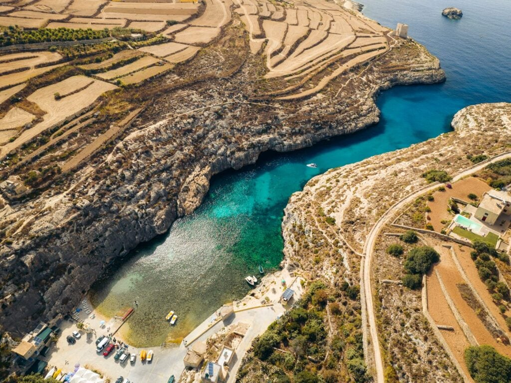 Aerial view of Mġarr ix-Xini Bay in Gozo, Malta, with terraced farmland leading to a secluded turquoise bay surrounded by rocky cliffs.