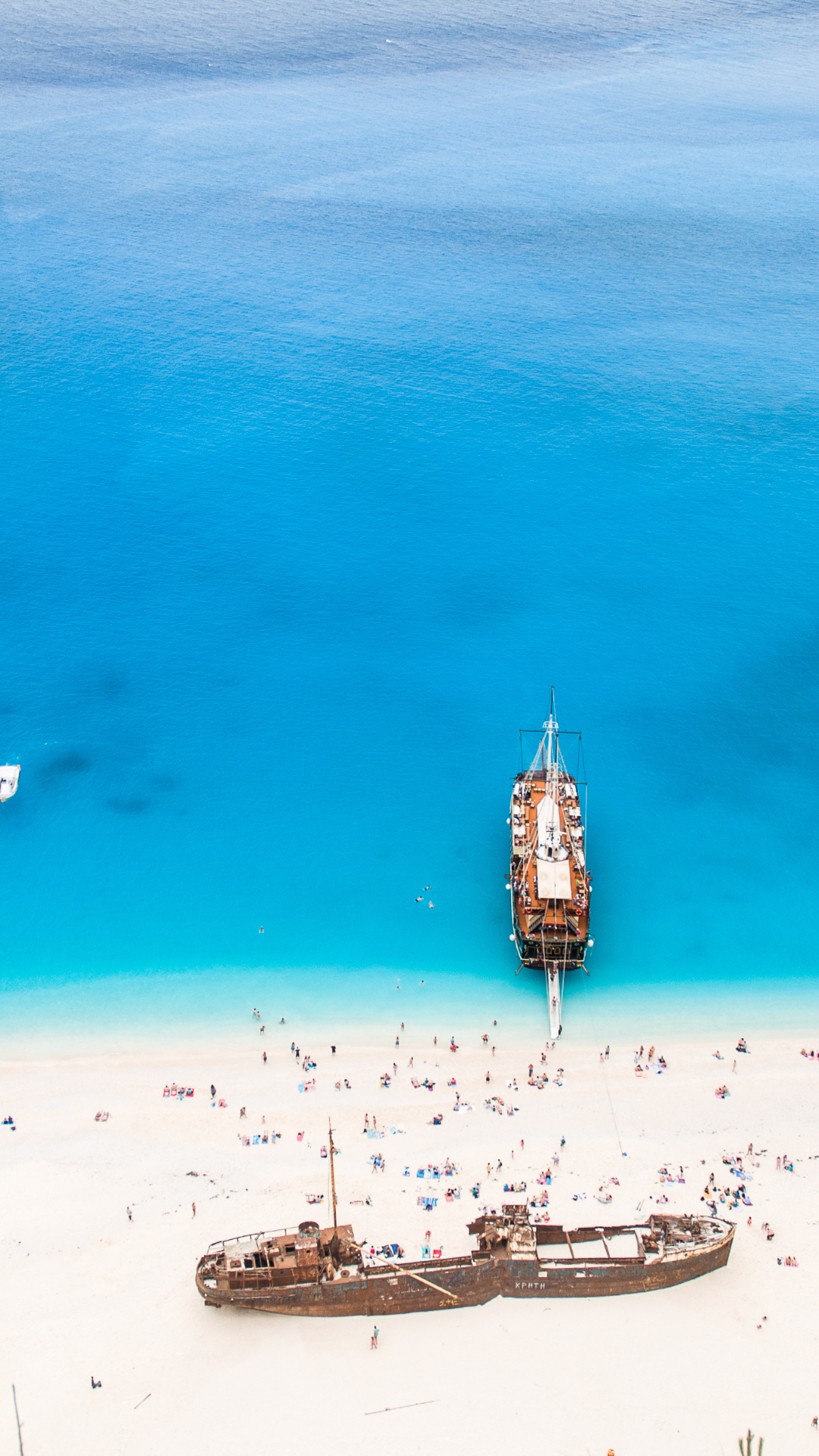 Beach goers at Navagio Beach hanging out by an old shipwreck on the beach with beautiful clear blue waters that has a large boat with people getting off to enjoy the boat.