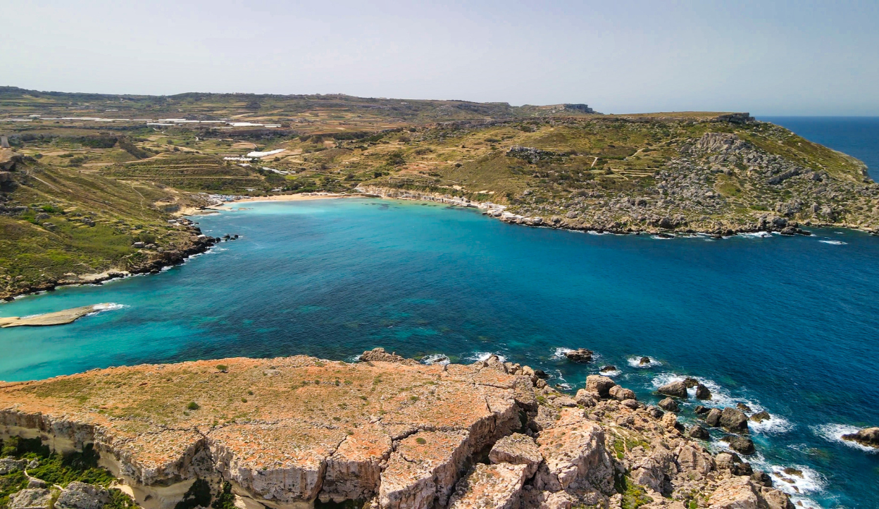 Aerial view of Paradise Bay in Malta, a secluded crescent-shaped beach with golden sand, clear turquoise waters, and rugged cliffs.