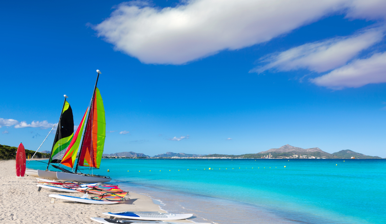 Colorful sailboats and paddleboards lined up on the soft white sand of Platja de Muro in Mallorca, Spain, with bright blue water and distant mountains