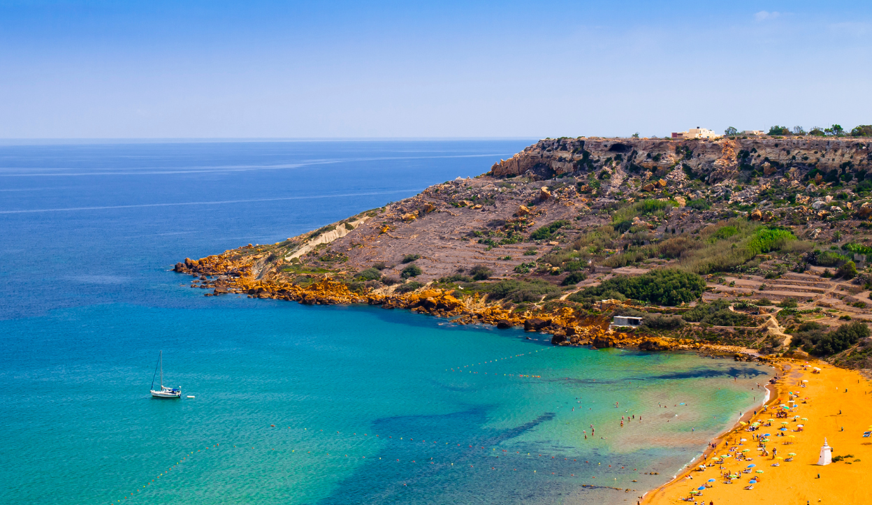 Scenic view of Ramla Bay in Gozo, Malta, with its unique red-golden sandy beach curving into turquoise waters and surrounded by rocky cliffs and farmland.