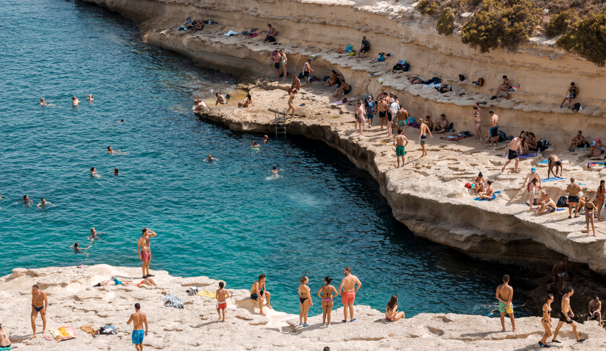 Crowded summer scene at St. Peter’s Pool in Malta, with people sunbathing on flat limestone cliffs and swimming in the bright blue Mediterranean waters.