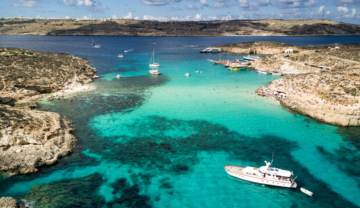 Aerial view of the Blue Lagoon in Comino, Malta, showing vivid turquoise waters, anchored boats, and swimmers between rocky islands.