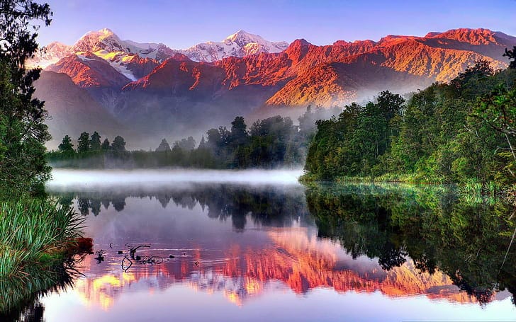 Still waters of Lake Matheson reflecting the Southern Alps at sunrise, surrounded by lush native forest in Westland Tai Poutini National Park, New Zealand.