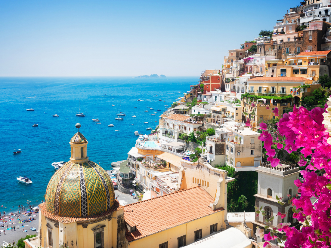 Seaside buildings in Naples Italy, with bright flowers seen besude white cliffside buildings over looking the sea below.