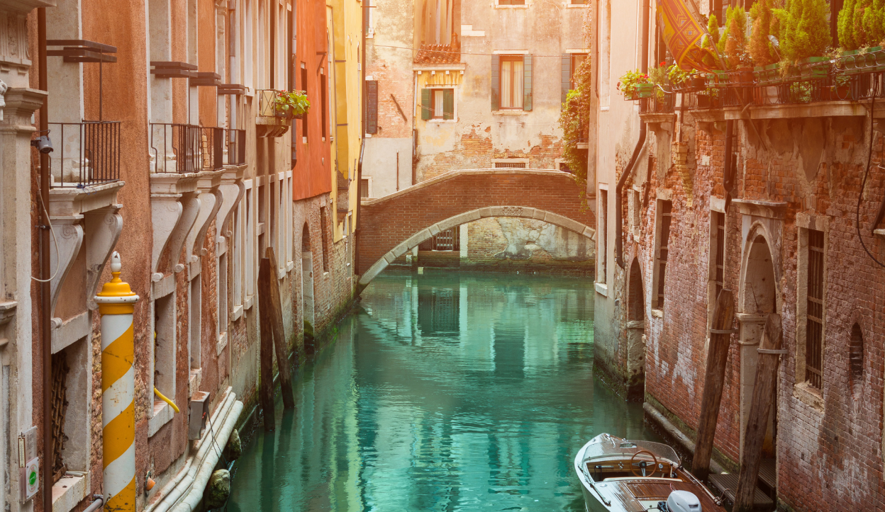 Venice, Italy canal between two buildings with a bridge connecting them, and a wooden boat tied to the building.