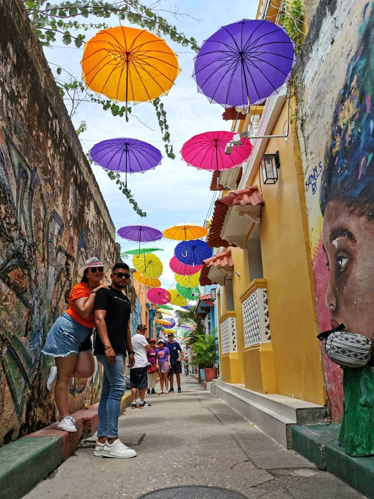 A narro street with Coloful umbrellas above visitors heads that go between each side of the narrow street to create head coverage.