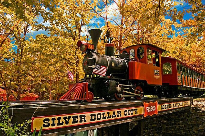 An 1800's style red train inside Silver Dollar City in Branson, with fall colored leaves on the Forrest trees behind it.