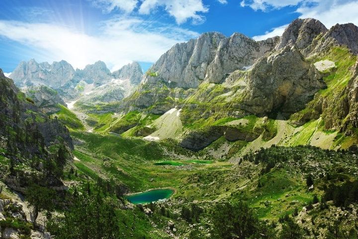 Theth National Park in Albania with dramatic peaks of the Accursed Mountains, alpine meadows, and glacial lakes under a bright blue sky.
