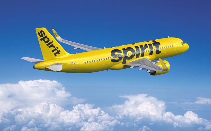 Spirit Airlines yellow jet flying in the sky with clouds below the jet. 