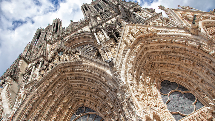 Close-up view of the ornate Gothic facade of Notre-Dame de Reims Cathedral in France, showcasing detailed stone carvings and arched entrances beneath a partly cloudy sky.