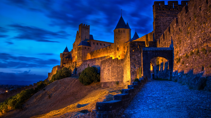 Night view of the medieval fortified city of Carcassonne in France, with its stone walls, watchtowers, and arched entrance illuminated by golden lights.