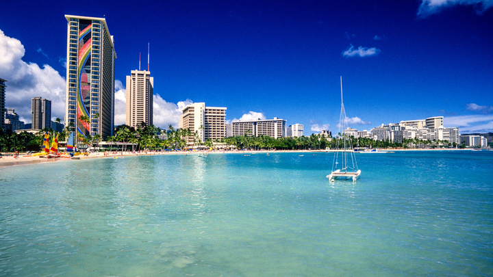 A catamaran anchored in clear blue waters off the beach in O'ahu, Hawaii with resorts lining the beach.