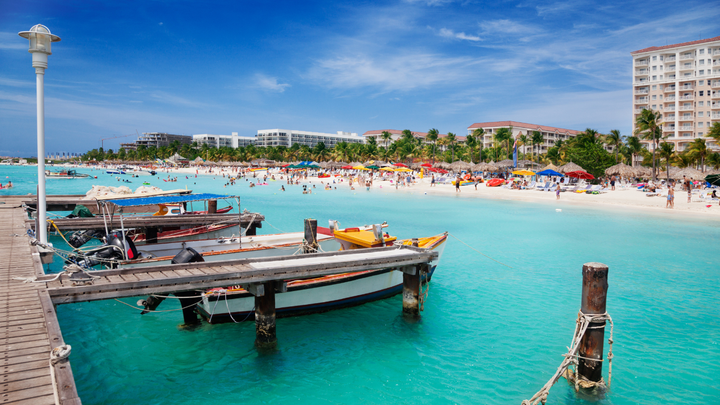 Aruba beach resorts with people swimming and playing on the beach and a boat dock with boats tied to it.