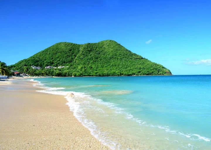A beautiful beach in St. Lucia with clear blue waters and a mountain covered in lush green trees beside the beach.