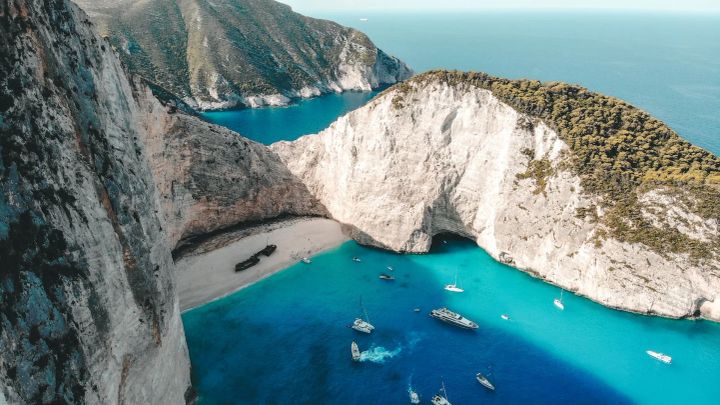Ariel view of a beach with a shipwreck on the beach, and sailboats anchored in the water near the beach at Navagio Beach in Greece.