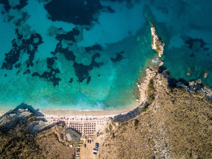 Aerial view of Mirror Beach (Pulebardha) in Albania with turquoise water, rocky coastline, and rows of umbrellas on the sandy shore.