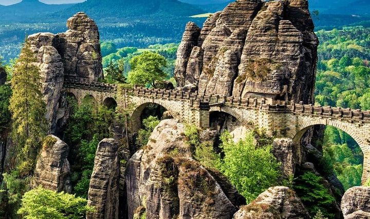 Historic Bastei Bridge spanning towering sandstone rock formations surrounded by lush green forest in Saxon Switzerland National Park, Germany, with distant hills visible in the background.