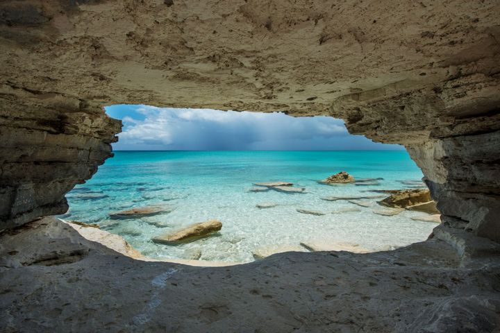 View from inside a rocky cave at Water Cay in Turks and Caicos, overlooking clear turquoise waters, scattered rocks in the shallow sea, and a dramatic sky with distant rain clouds.