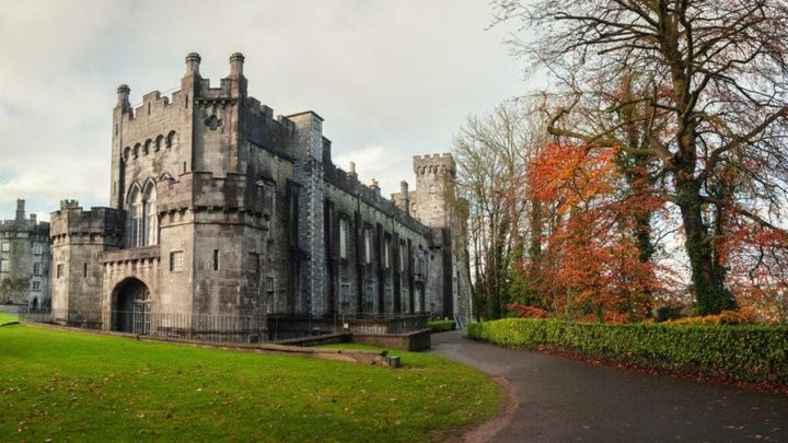 Historic Kilkenny Castle in Ireland surrounded by lush green lawns and vibrant autumn trees with orange and red foliage, under a cloudy sky.