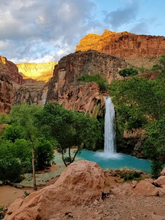 Havasu Falls is a water fall that comes out of red rocks in a canyon and falling into a clear blue pool of water.