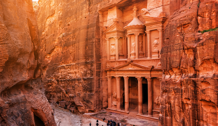 Al-Khazneh, the carved sandstone Treasury in Petra, Jordan, framed by towering red rock cliffs with tourists gathered below.