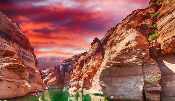 Kayakers paddle through Antelope Canyon at Lake Powell, with towering red rocks under a vibrant red and purple sunset sky.