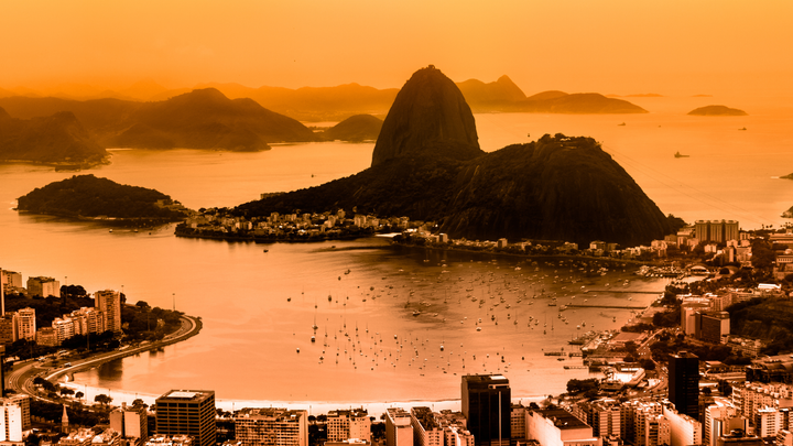 Aerial view of Rio de Janeiro, Brazil, showcasing Sugarloaf Mountain, Guanabara Bay filled with boats, and the cityscape at sunset with an orange hue.