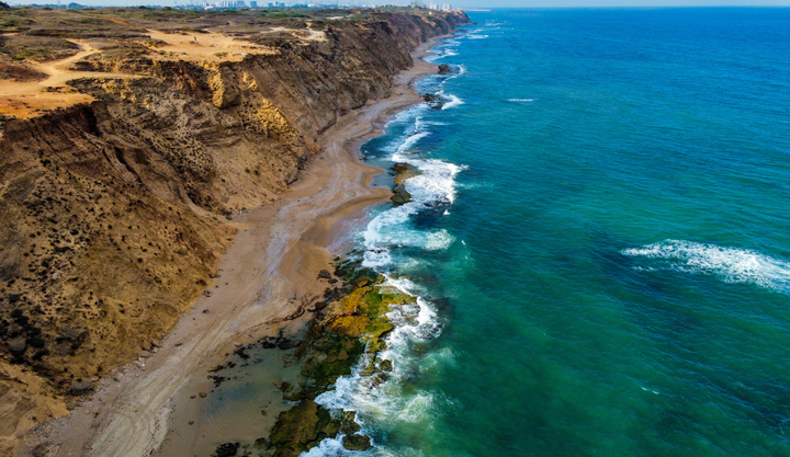 Waves crashing on the shore of Arsuf Beach in Israel with large cliff behind the beach and clear emerald waters.