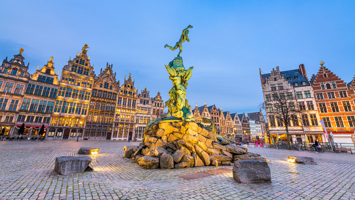 Brabo Fountain in Grote Markt square, Antwerp, Belgium, with ornate guildhall buildings lit at dusk under a clear blue sky.