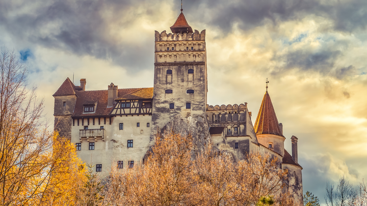Bran Castle, also known as Dracula’s Castle, perched atop a rocky hill and surrounded by autumn trees under a dramatic, cloudy sky in Transylvania, Romania.