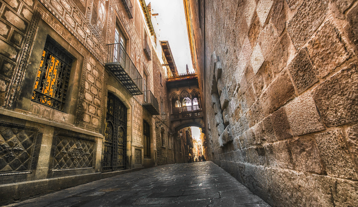 Stone alleyway in Barcelona’s Gothic Quarter featuring medieval buildings and the ornate Pont del Bisbe bridge overhead.