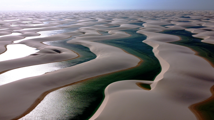 Aerial view of Lençóis Maranhenses National Park in Brazil, showing endless white sand dunes interspersed with turquoise rainwater lagoons under a clear sky.