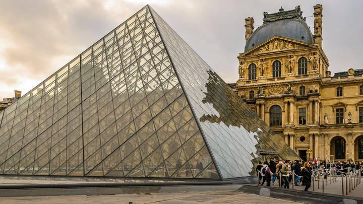Visitors line up outside the glass pyramid entrance of the Louvre Museum in Paris, with the historic palace building in the background under a cloudy sky.