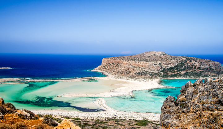 A panoramic view of Balos Beach in Crete, Greece, featuring turquoise shallow lagoons, white sandy shores, and the rocky Gramvousa Peninsula under a clear blue sky.
