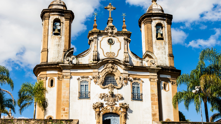 Baroque-style colonial church with twin bell towers in Ouro Preto, Brazil, set against a blue sky and surrounded by palm trees.