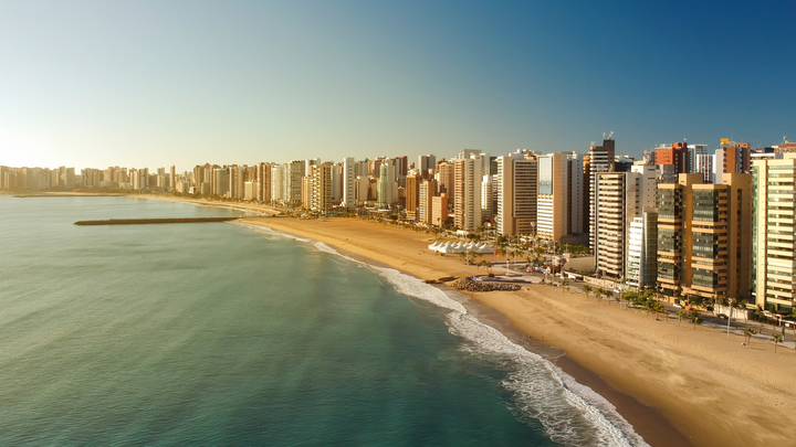 Aerial view of beachfront resorts and high-rise buildings lining the sandy shore in Fortaleza, Brazil, with calm ocean waves and a clear sky.