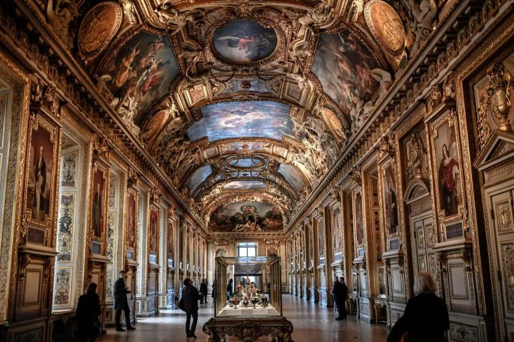 Visitors walk through an ornate gallery in the Louvre Museum, Paris, with gilded walls, framed portraits, and a vaulted ceiling adorned with elaborate paintings.