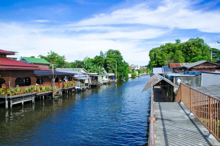 Scenic view of traditional wooden stilt houses lining a peaceful canal at Khlong Bang Luang Floating Market in Bangkok, under a bright blue sky.