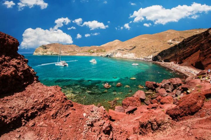 A panoramic view of Red Beach in Santorini, Greece, featuring vibrant red volcanic cliffs, turquoise waters, anchored sailboats, and beachgoers sunbathing along the narrow shoreline.