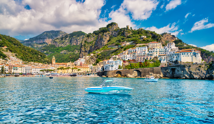 Scenic view of the Amalfi Coast in Italy, featuring colorful hillside buildings, lush green mountains, and boats floating on the clear blue waters of the Mediterranean Sea.